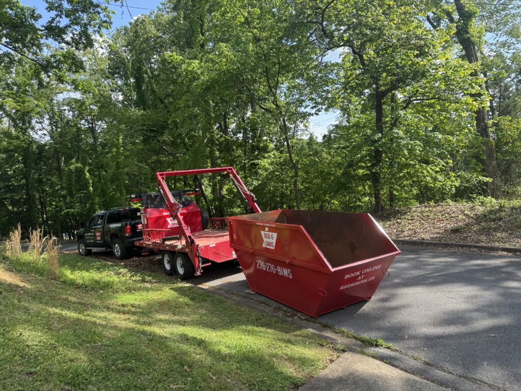 Red-E-Bins dumpster rental setup in a scenic, tree-lined neighborhood. A black pickup truck is towing a bright red dumpster trailer with a 15-yard Red-E-Bins dumpster placed on the roadside. The dumpster is labeled with the Red-E-Bins logo, contact number, and website for easy booking. The surroundings feature lush green trees and a paved road, showcasing the clean and driveway-safe placement of the dumpster.